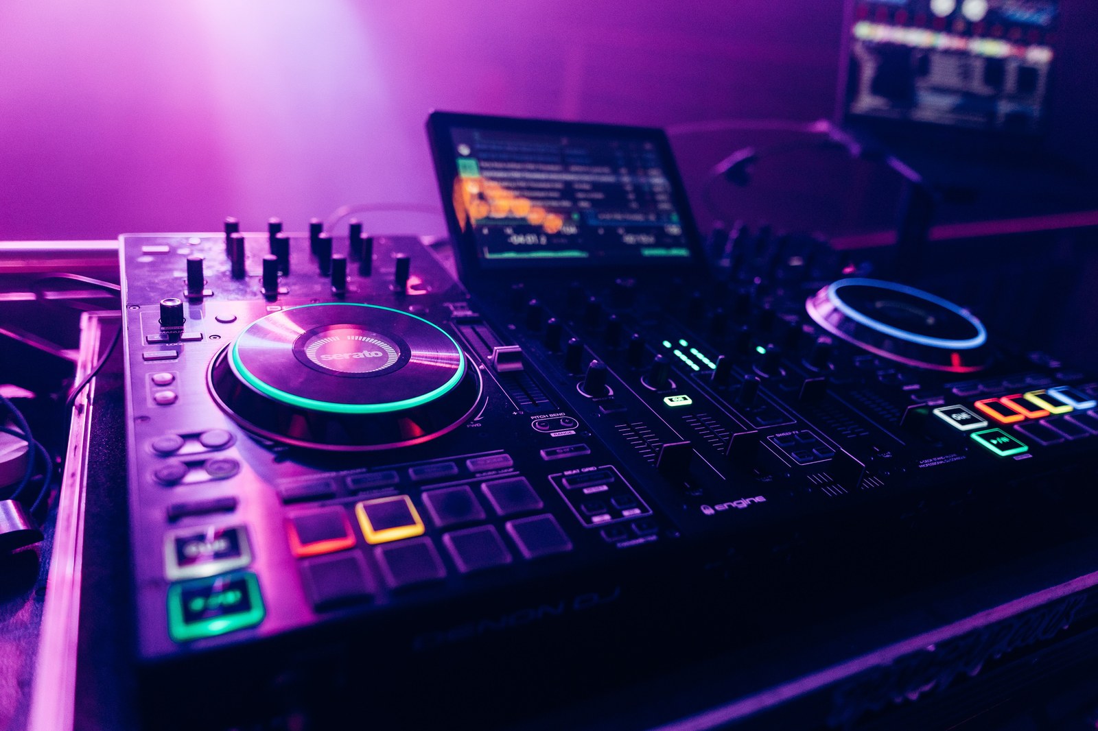 A DJ controller and mixer glowing in purple and blue club lighting, with a jog wheel lit green and a laptop screen showing a track in the background
