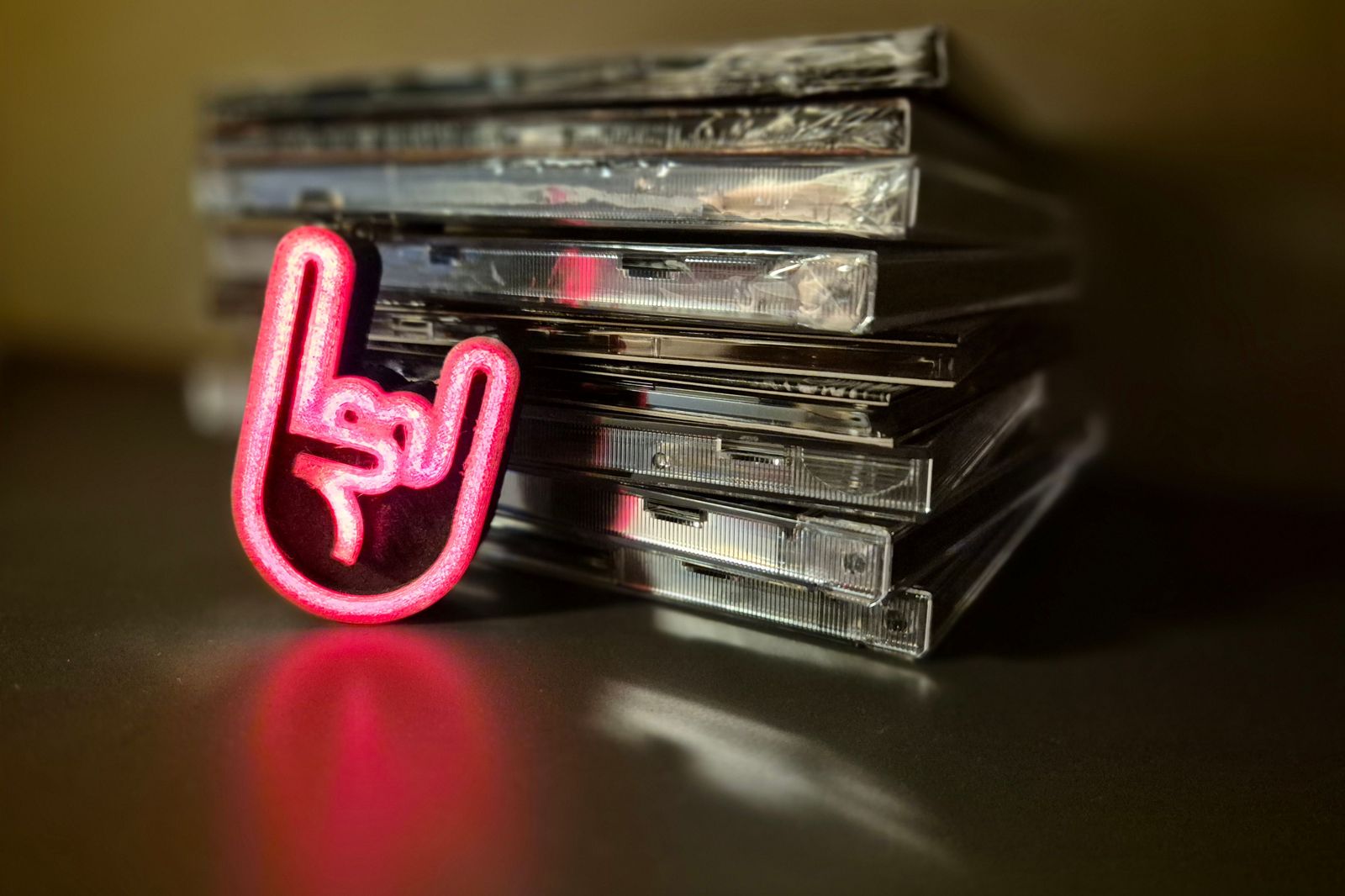 A glowing pink rock-hand symbol leaning against a stack of CD cases on a dark, reflective surface